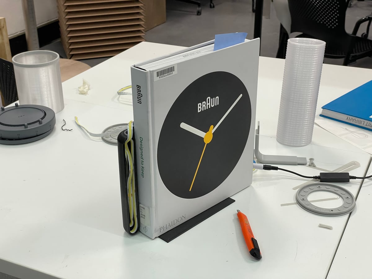 Workbench shot of a Braun alarm clock beside a ribbed white cylinder prototype, 3D-printed rings, and other parts