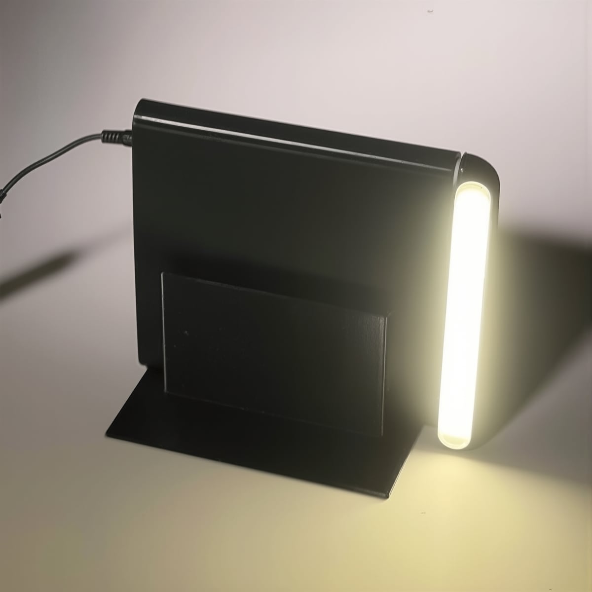 Phone photo of a black prototype bookend with a glowing white light strip and cable on a gray base plate against a pale wall