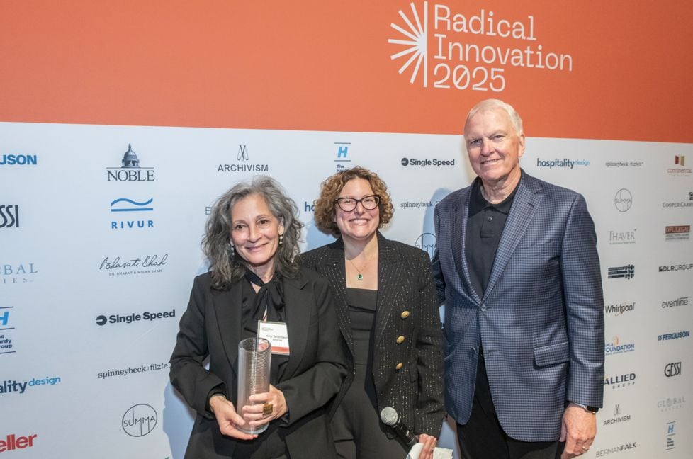 Event photo of three people (two women, one older man in blazer) posing in front of the orange Radical Innovation 2025 sponsor backdrop