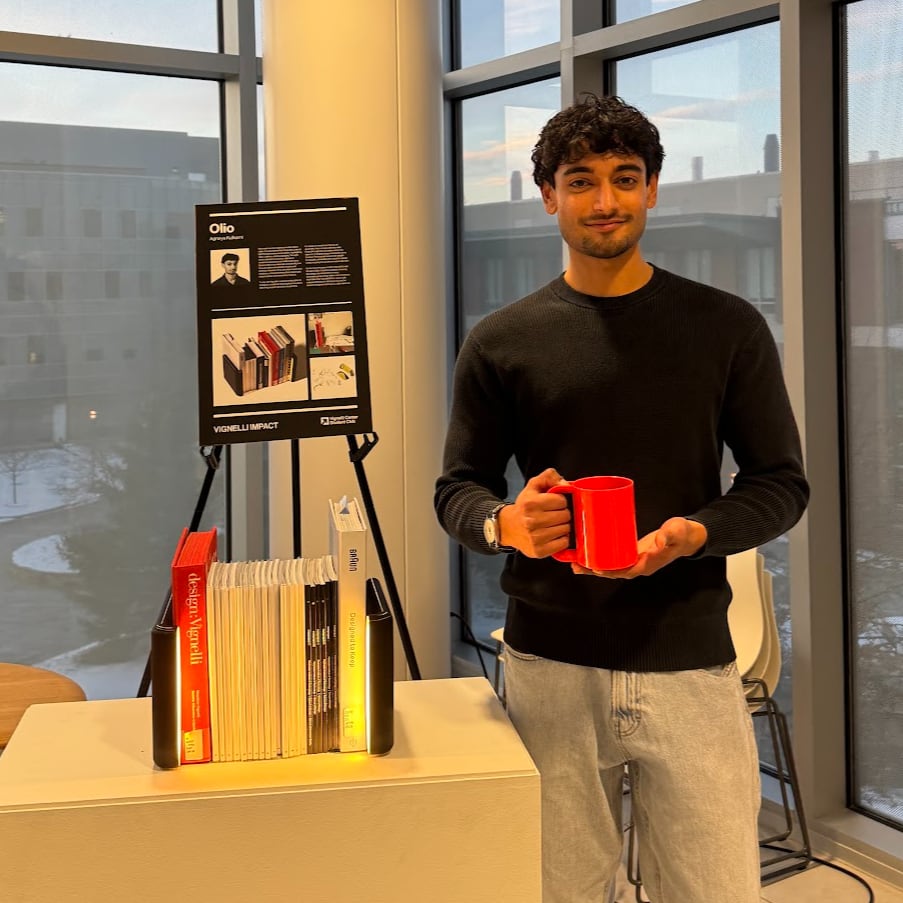 Young man in a black sweater holding a red mug beside a display stand with a poster and a bookend with books on a white plinth, in front of large