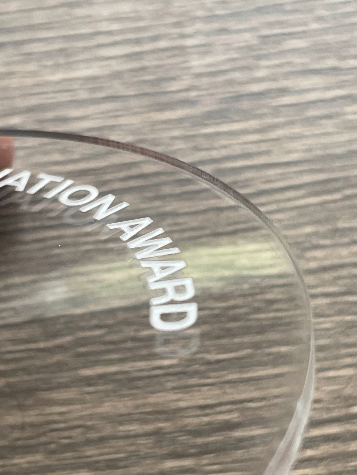 Close-up of clear acrylic disc embossed with partial text 'ATION AWARD' resting on a wood surface